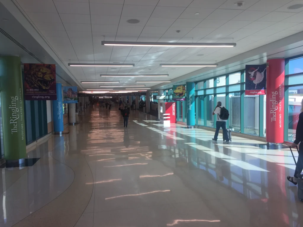 Wide airport corridor with colorful advertisement banners, several people walking, sunlight streaming through large windows on the right side.