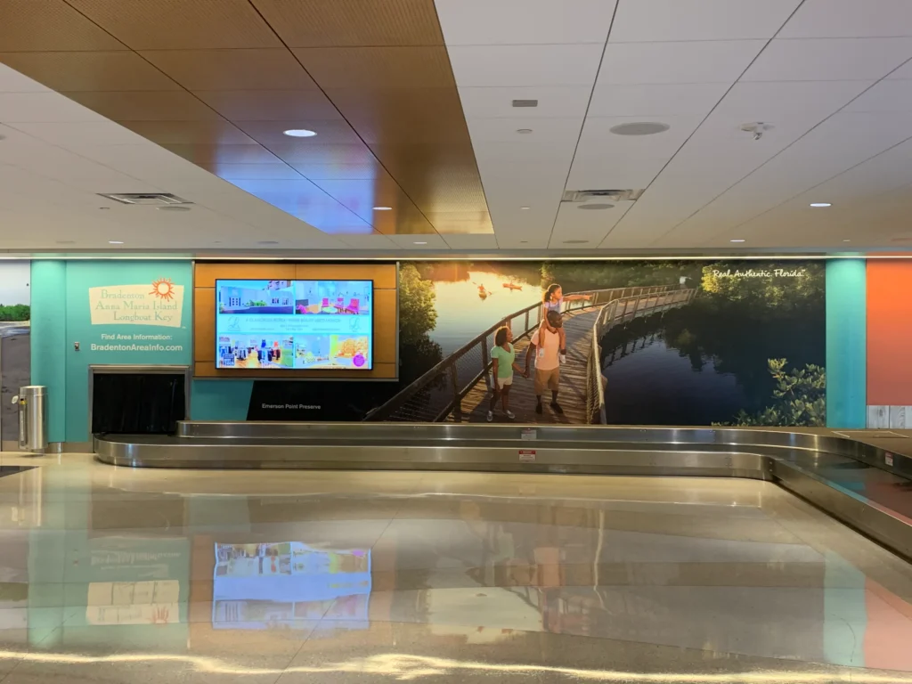 Baggage claim area with a Bradenton area tourism ad, digital display, and a large mural of a family walking on a boardwalk over water.