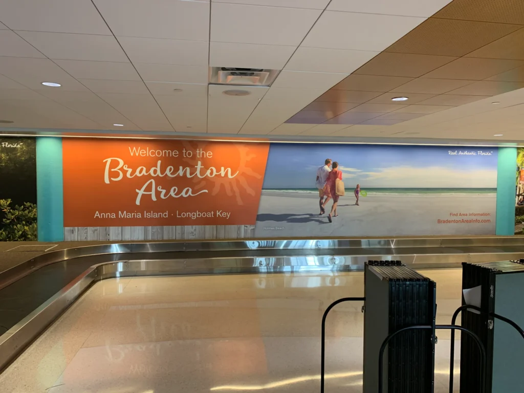 Large airport wall ad reads “Welcome to the Bradenton Area, Anna Maria Island - Longboat Key” with a photo of a family walking on a sunny beach by the sea.