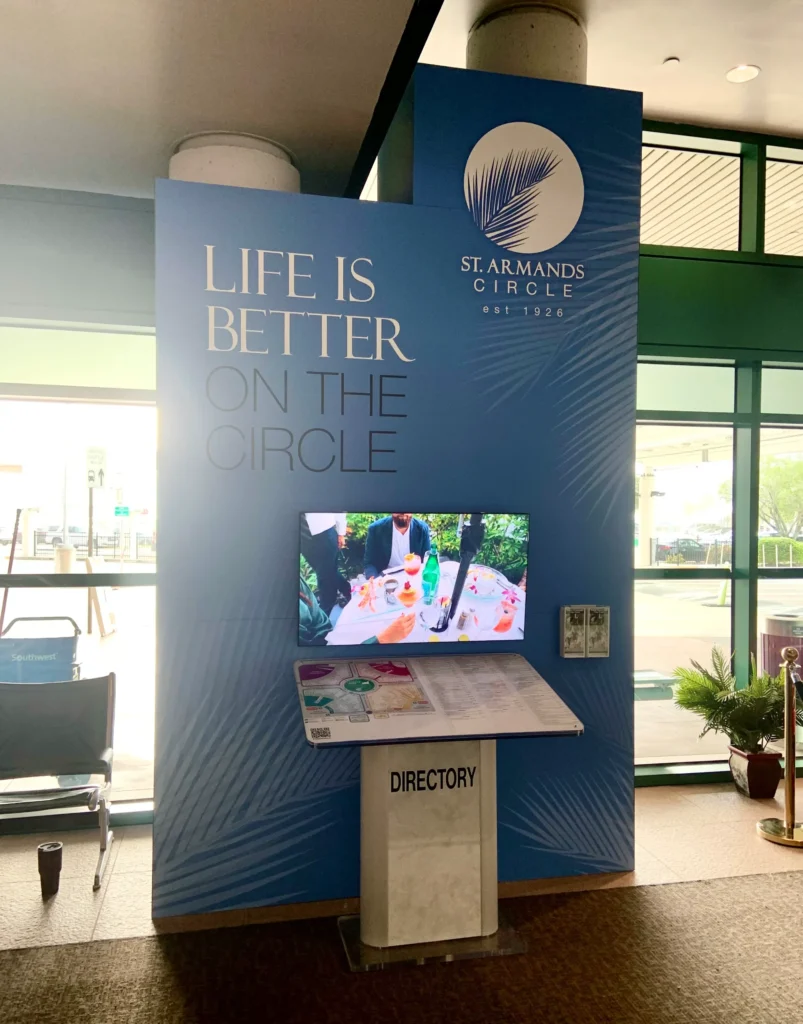 A St. Armands Circle display with a slogan, a digital screen showing people at a table, and a directory stand in front.