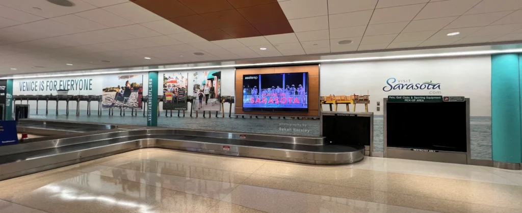 Baggage claim area with a Sarasota promotional wall display, digital screen, and luggage conveyor belt in an airport.