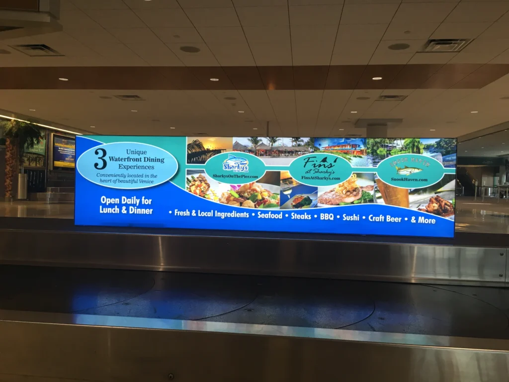 Billboard at baggage claim advertises three waterfront dining spots, featuring seafood, BBQ, sushi, steaks, craft beer, and more. Banners show restaurant logos and food images.