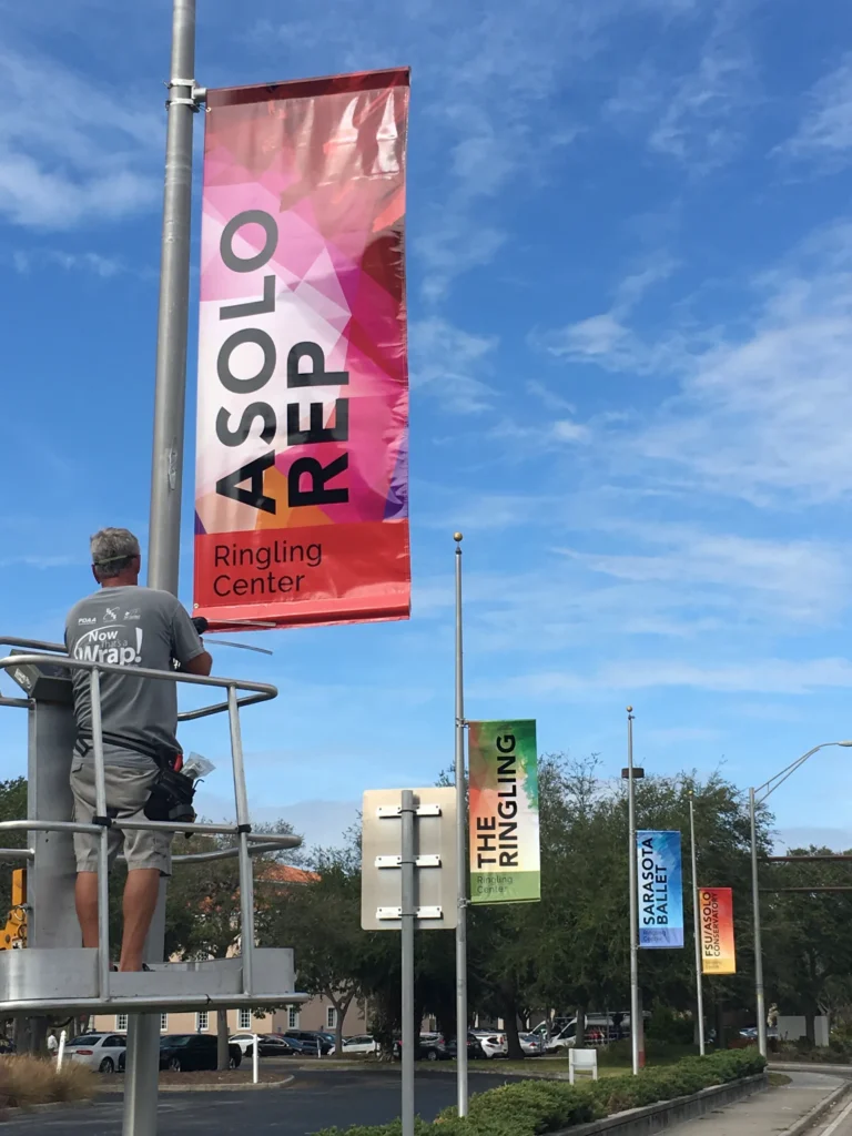 A man in grey stands on a lift adjusting a red “Asolo Rep Ringling Center” banner; other colorful banners and trees line the street under a blue sky.