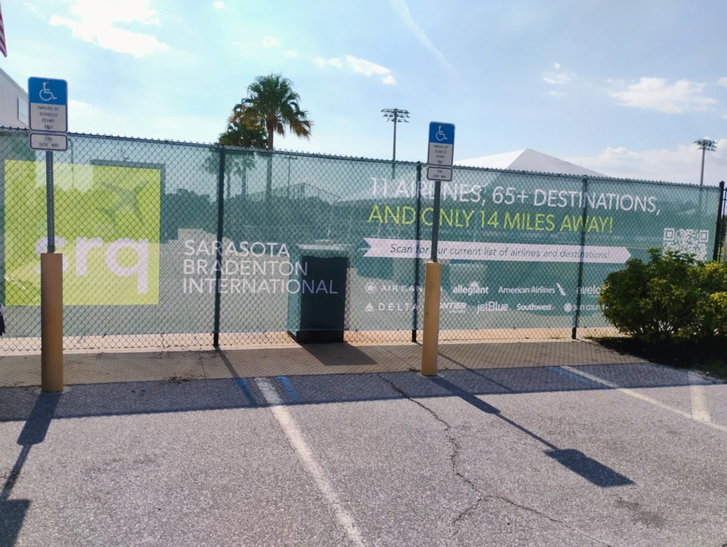 Green and white fence sign for Sarasota Bradenton International Airport promotes 11 airlines and 65+ destinations; parking spaces with handicap signs are visible in front.