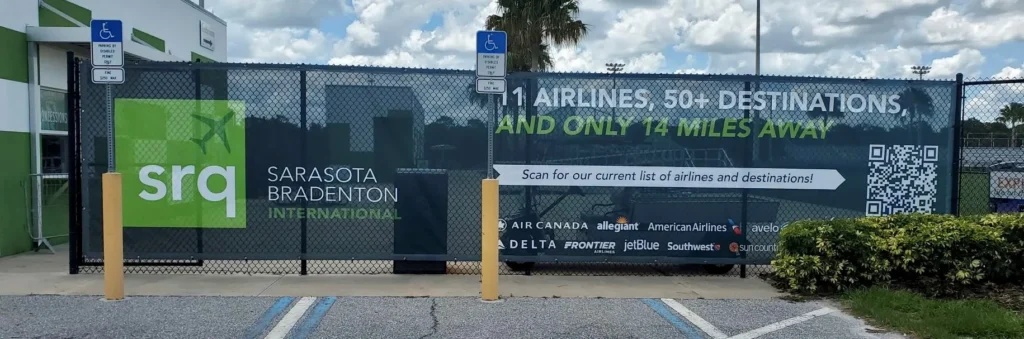 Banner at Sarasota Bradenton International Airport advertises 11 airlines, 50+ destinations, and a QR code for flight info; two handicapped parking signs are visible in front of the fence.