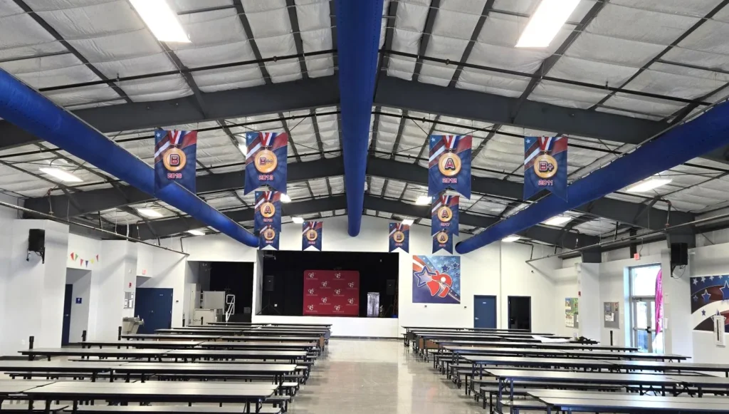 School cafeteria with long tables, blue ventilation pipes on the ceiling, banners hanging, and a stage at the far end.