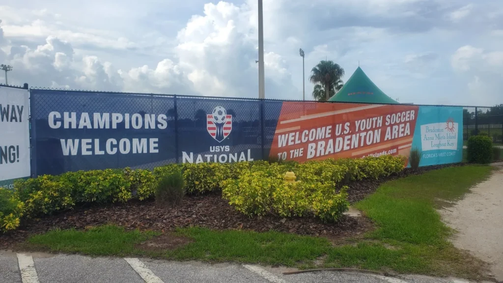 Banners on a fence read “Champions Welcome,” “USYS National,” and “Welcome U.S. Youth Soccer to the Bradenton Area” beside landscaping and a green tent.