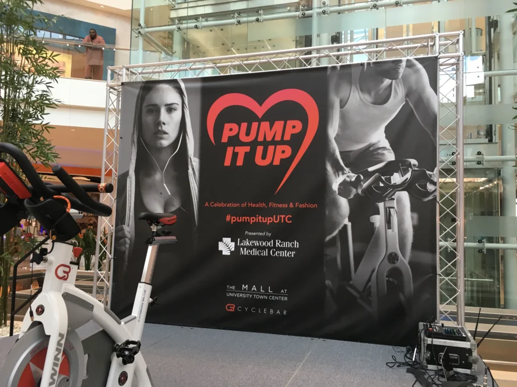 A stage backdrop displays “PUMP IT UP” and event details for a fitness celebration at The Mall at University Town Center, with images of a woman and a person cycling. A stationary bike is in front.