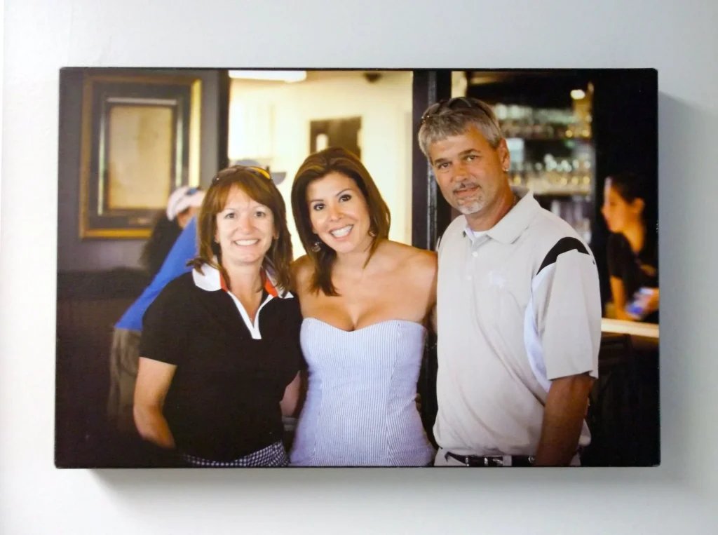 Three adults stand together indoors, smiling at the camera; a woman in a white dress is between a man and a woman in casual clothes.