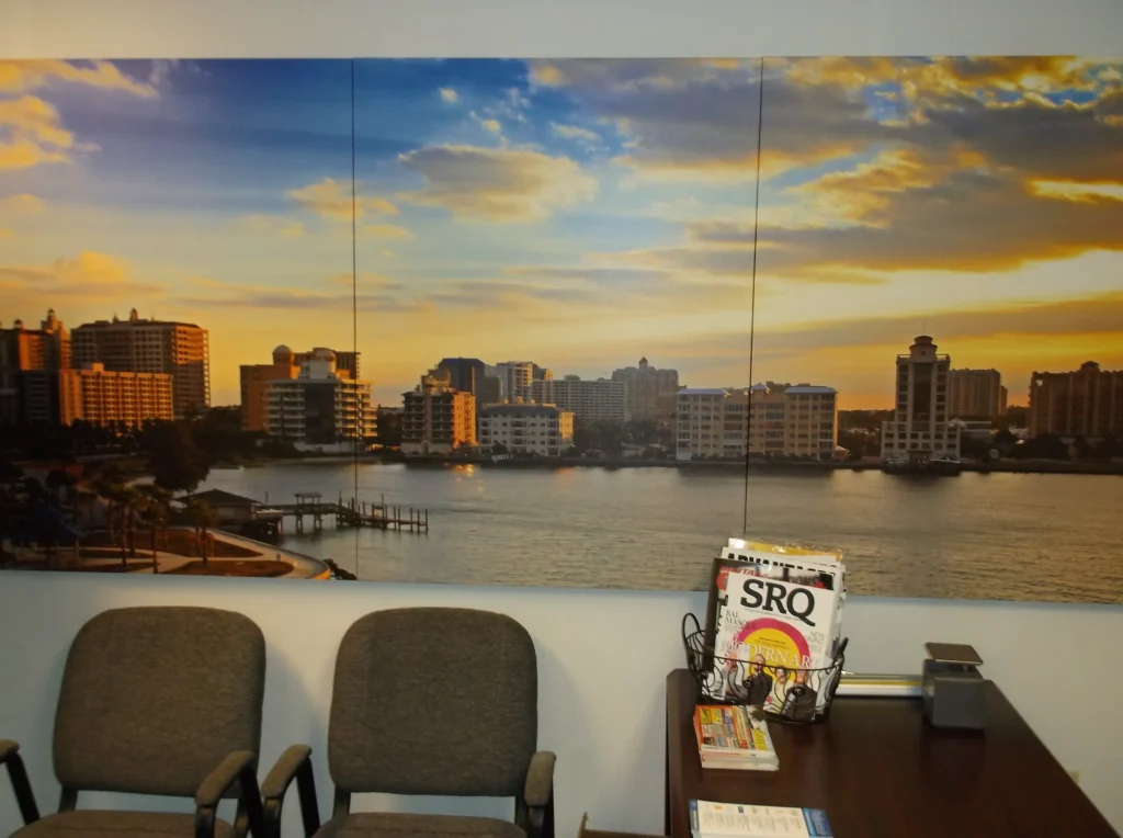 Large wall art of a cityscape over water at sunset hangs above gray chairs and a table with magazines and brochures.