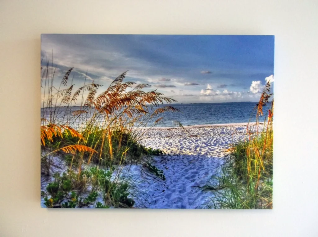 A canvas print of a sandy beach pathway with grass leads to the ocean under a blue sky with clouds.