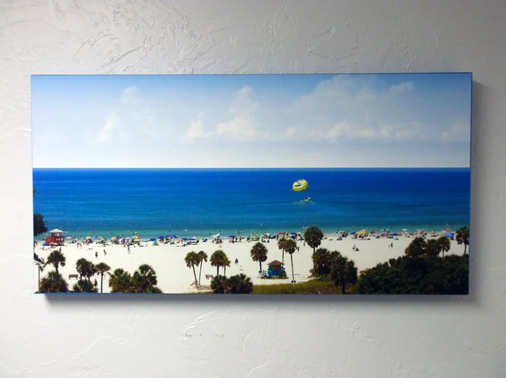 A framed photo of a beach with white sand, palm trees, blue ocean, people sunbathing, and a parasailor in the sky is hanging on a textured white wall.