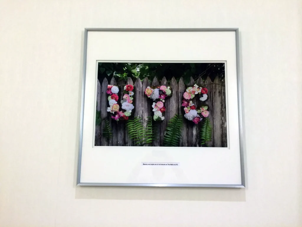 Framed photo of a wooden fence decorated with colorful flower arrangements and green ferns, hung on a plain white wall.