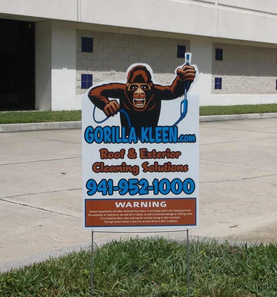 A yard sign for Gorilla Kleen advertises roof and exterior cleaning solutions with a cartoon gorilla, phone number, and website, placed on grass by a sidewalk.