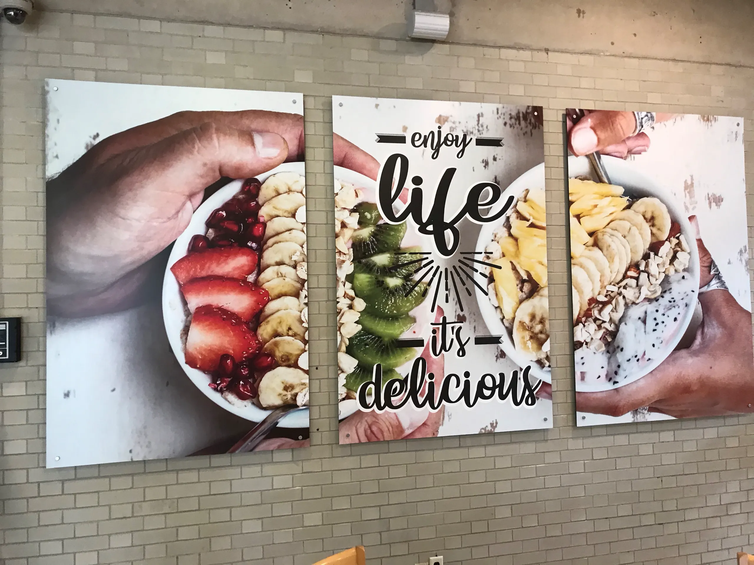 Three wall posters show hands holding smoothie bowls with fruit toppings and granola; the center poster reads “enjoy life it’s delicious” in bold, decorative text.