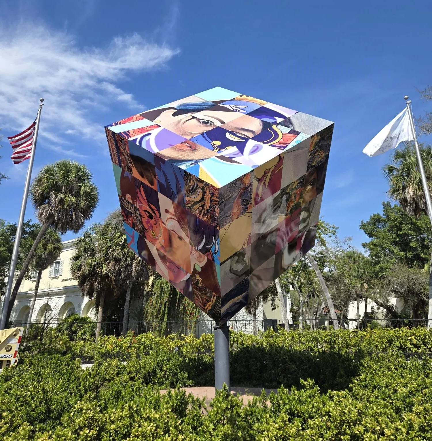 Large cube sculpture covered in colorful murals is mounted on a pole in an outdoor garden with palm trees and flags, under a clear blue sky.