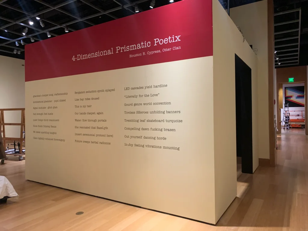 A beige exhibit structure with a red top panel titled “4-Dimensional Prismatic Poetix,” featuring poems by B. Cypress and Osceola Clan, displayed on the gallery’s wooden floor.