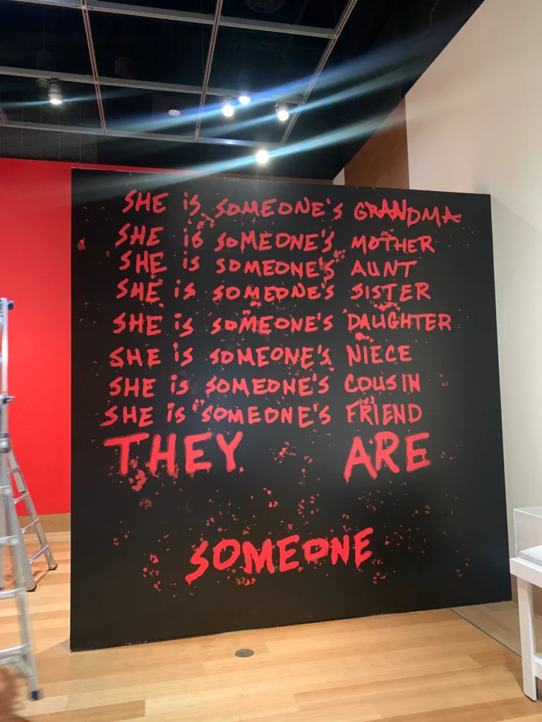 A black wall with red text listing relationships like grandma, mother, aunt, daughter, and ending with “THEY ARE SOMEONE.” A ladder is on the left and a white chair is on the right.