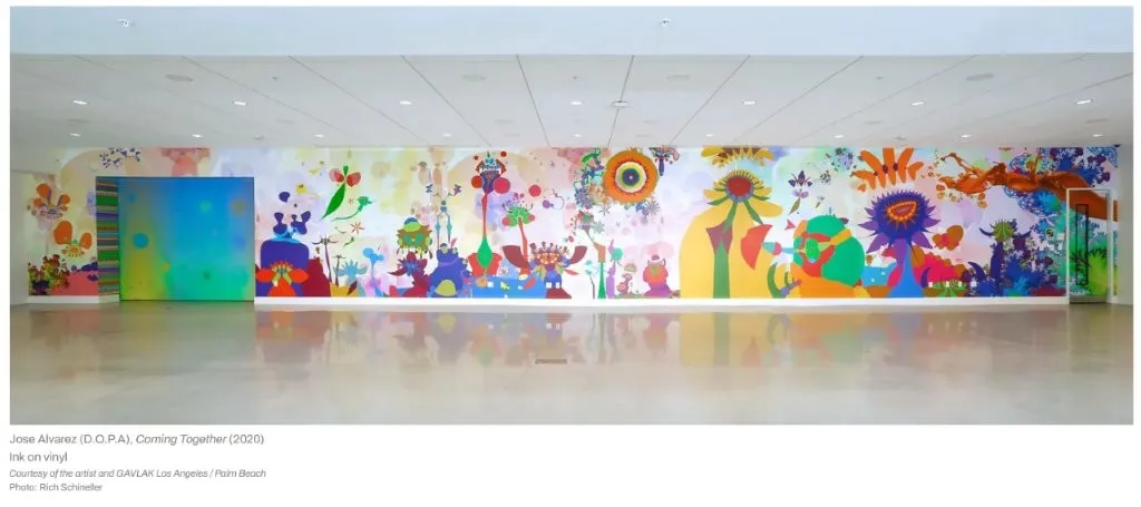 A vibrant mural with colorful, abstract plant and flower shapes stretches along a white wall in a spacious, modern gallery.