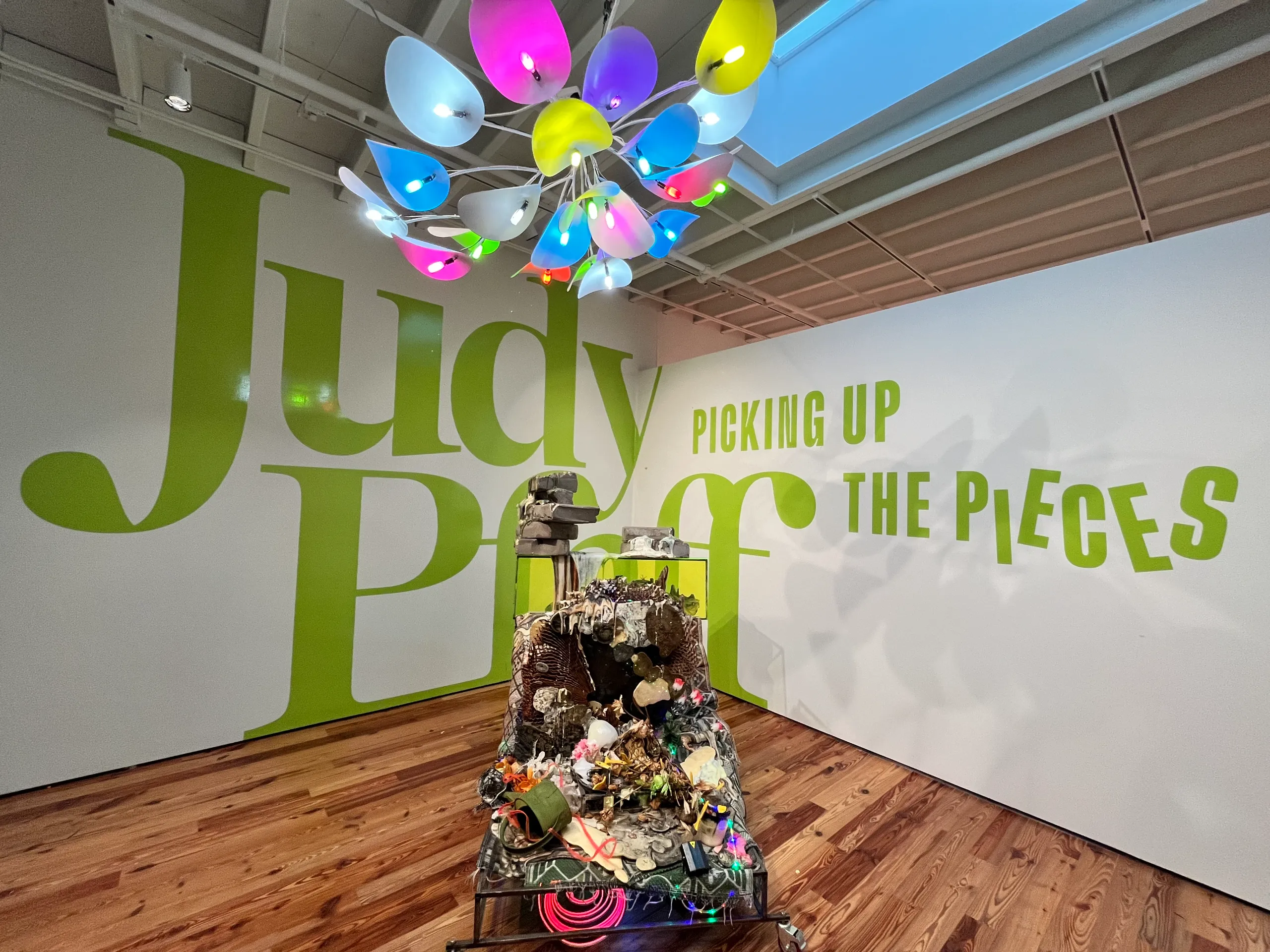 Art exhibit featuring a large “Judy Pfaff: Picking Up the Pieces” wall graphic, colorful ceiling lights, and a mixed-media sculpture on a wooden floor.