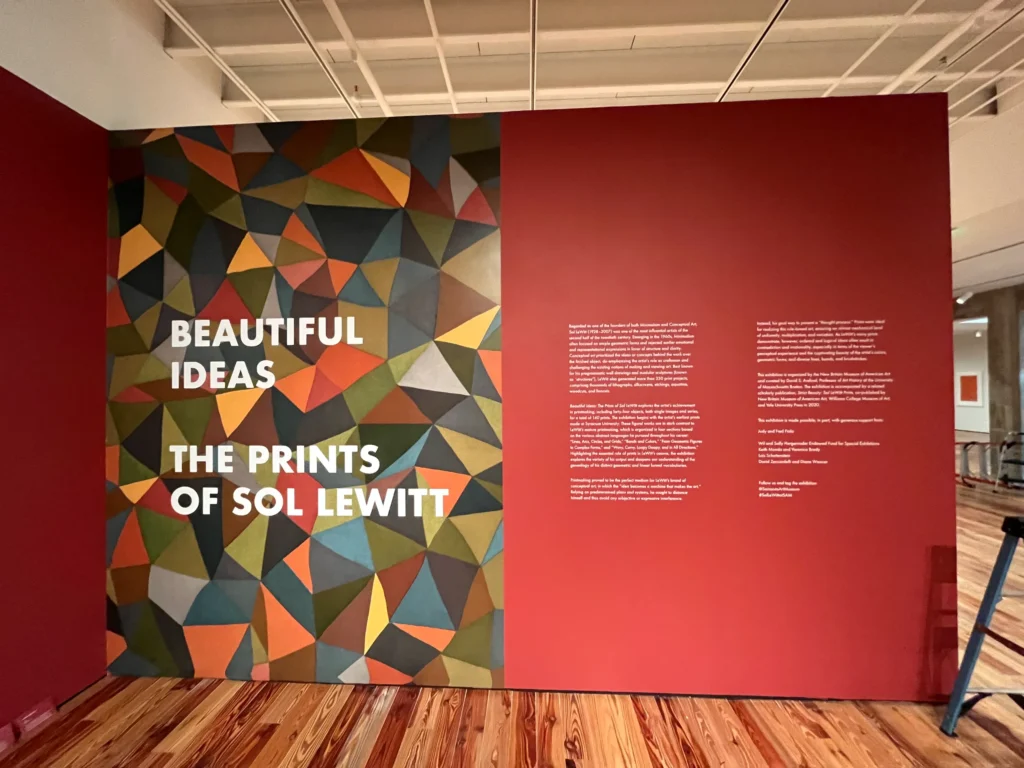 Exhibit wall with geometric pattern, large text reading “Beautiful Ideas: The Prints of Sol LeWitt,” and curatorial information on a red background.