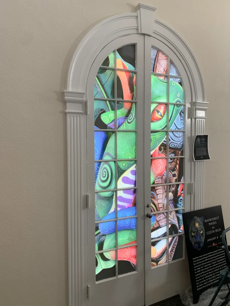A colorful projection of rainforest animal masks is displayed on glass-paneled double doors, with a nearby sign reading “Rainforest Masks, Costa Rica” and exhibit information.