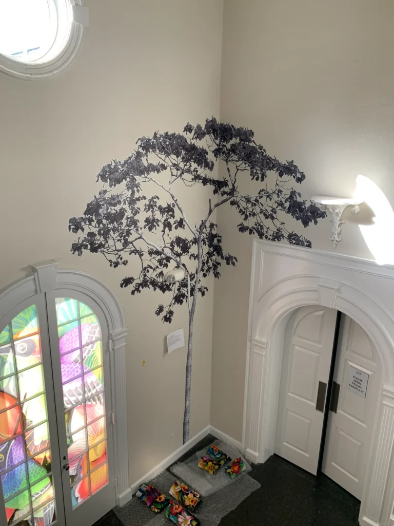 A black-and-white tree mural decorates a white wall above a mat with colorful shoes, near arched double doors and stained glass windows.