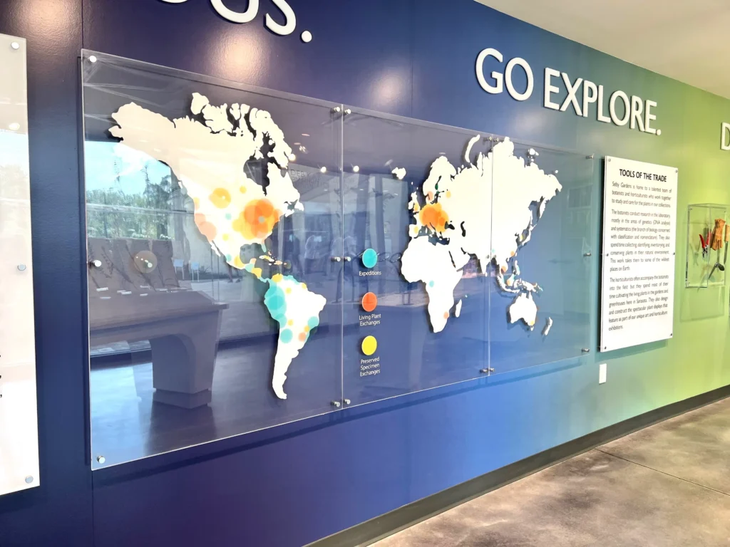 A glass world map with colored dots highlights ocean expeditions, ship locations, and coral reef sites, mounted on a blue wall in a museum or educational center.