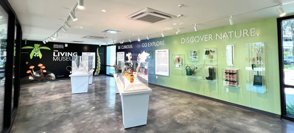 A modern museum exhibit room features plant displays, informational panels, and text on the wall reading “BE CURIOUS. GO EXPLORE. DISCOVER NATURE.”.