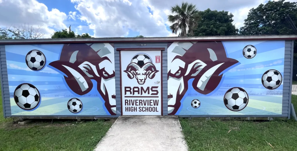 A building with a mural of two ram heads, soccer balls, and the text “Rams Riverview High School” above a door, set against a blue sky with clouds.