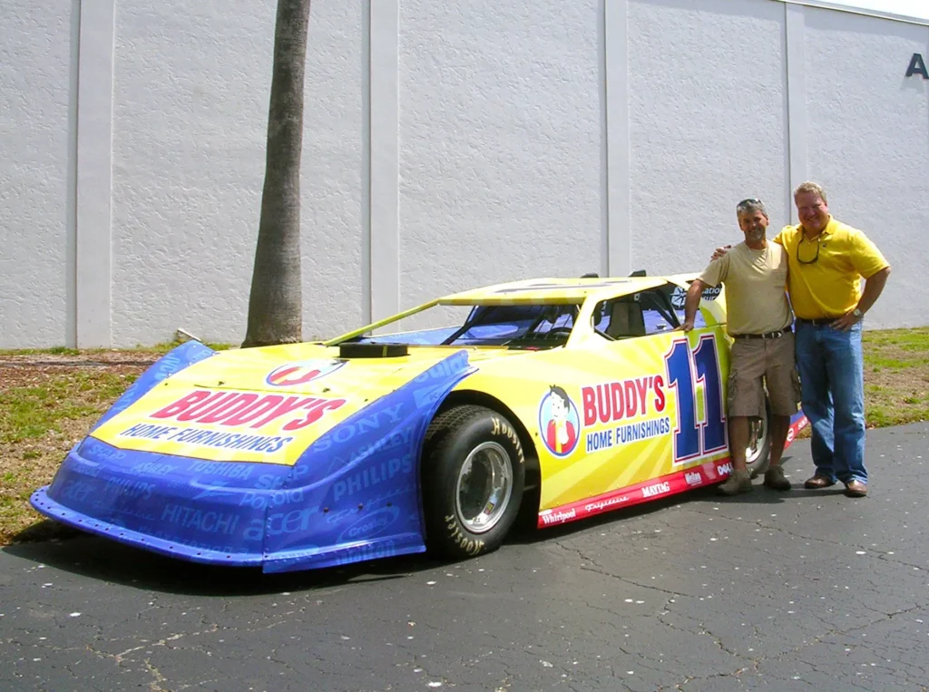 Two men stand beside a yellow and blue Buddy’s Home Furnishings race car parked on asphalt in front of a white building.