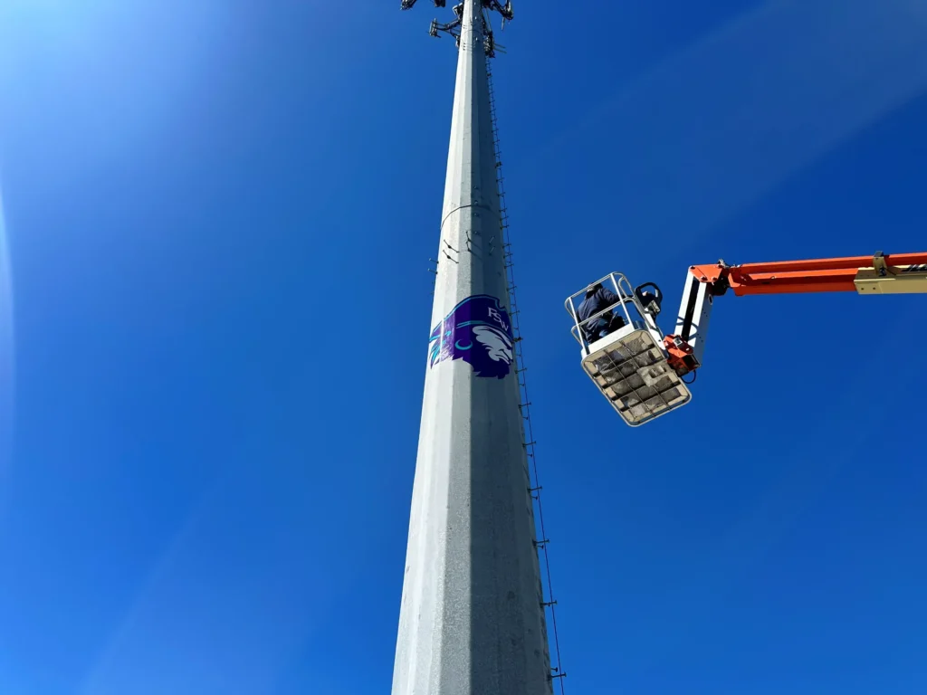 A person in a raised lift applies a purple Wildcat logo decal to a tall light pole against a clear blue sky.
