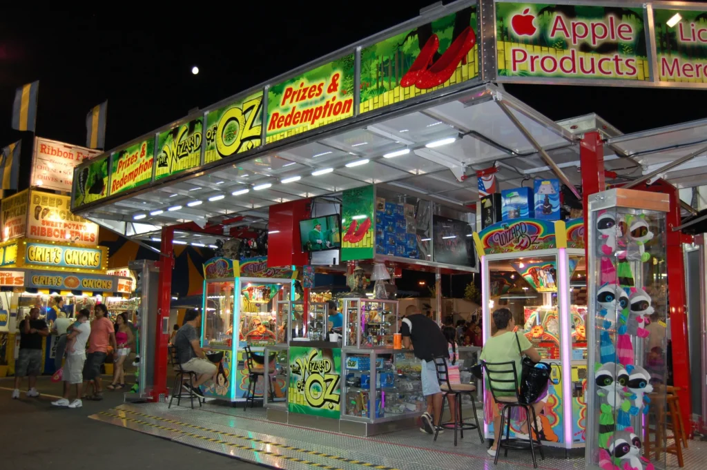 Carnival game booth at night with colorful signs, arcade machines, and people sitting on stools. Prizes and Apple products are advertised. Other booths and lights are visible in the background.