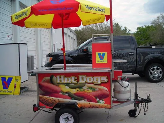 Hot dog cart with a Vienna Beef umbrella, propane tank, and hot dog graphics, parked near a black pickup truck outside a building.
