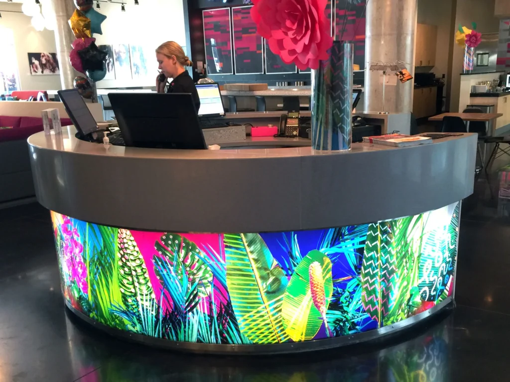 A woman works at a curved reception desk with colorful tropical leaf designs on the front in a modern office lobby.
