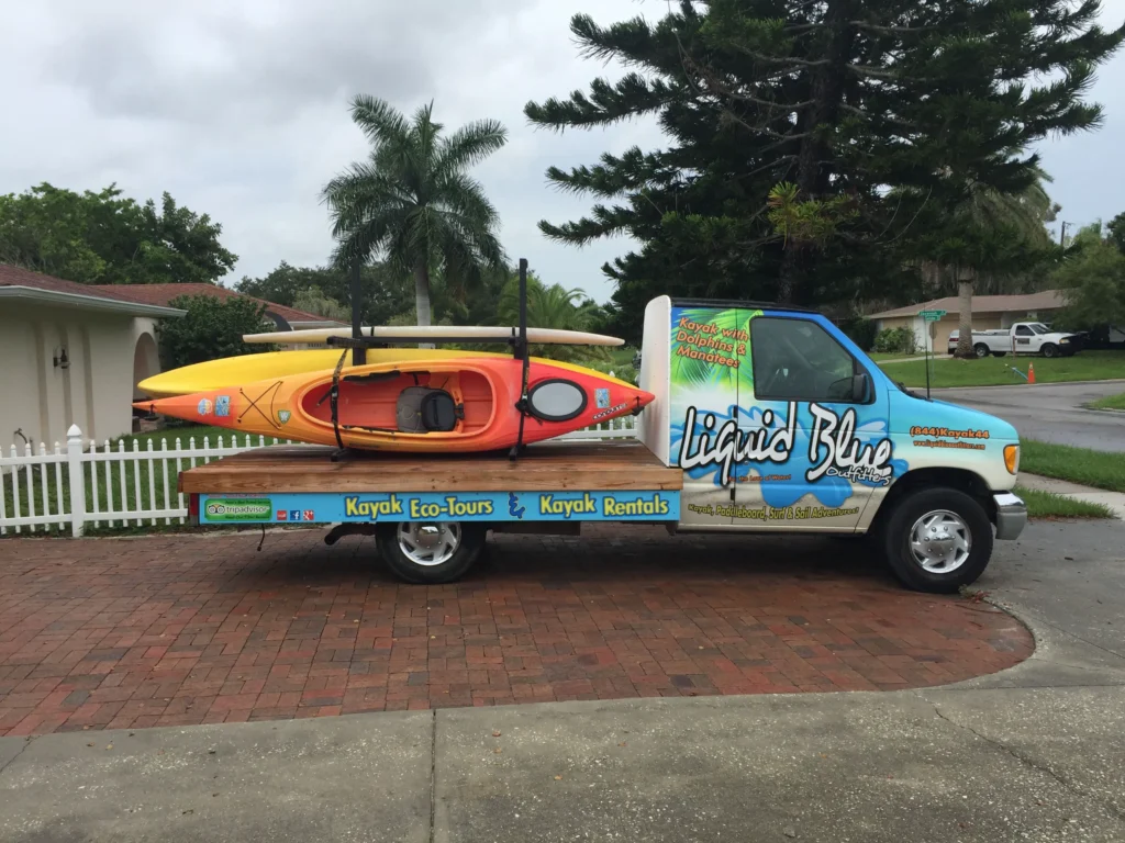 A flatbed van labeled “Liquid Blue” is parked on a driveway, carrying two kayaks stacked on its bed.