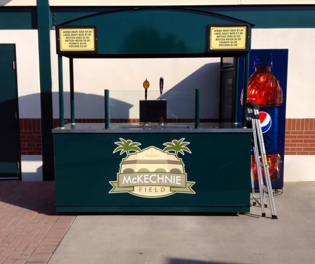 Green concession stand labeled “McKechnie Field” with a menu above, a large Pepsi bottle display on the right, and a folded silver ladder beside the counter.