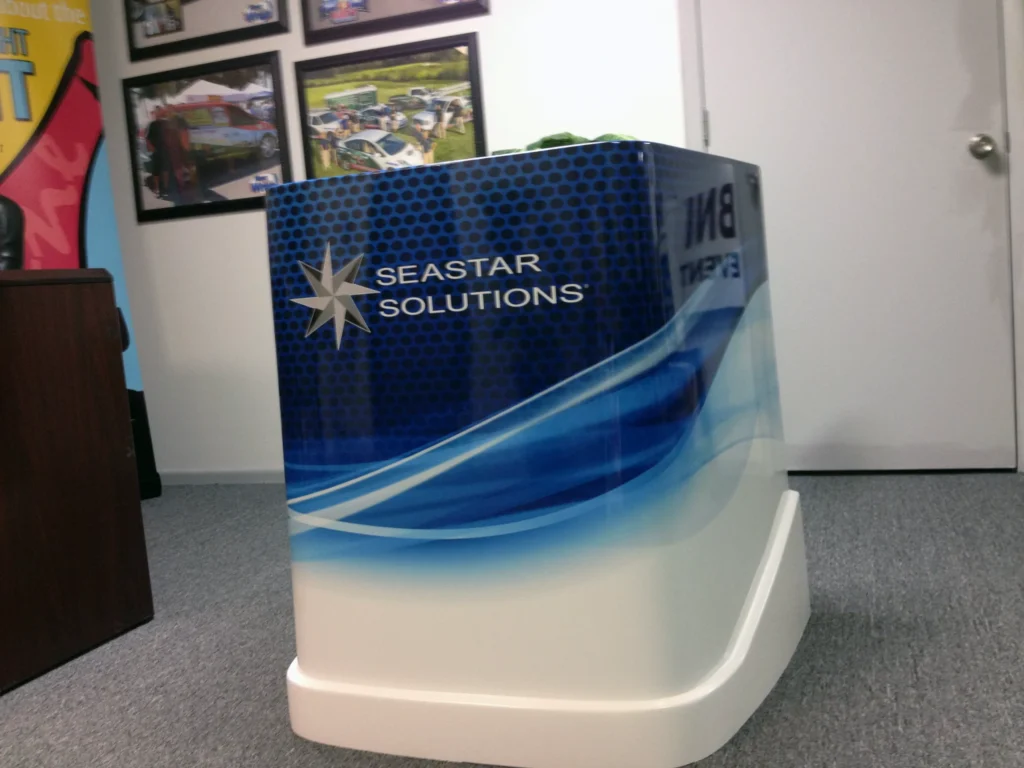A rectangular display stand with a blue and white design features the Seastar Solutions logo and name in an office setting with framed pictures on the wall.