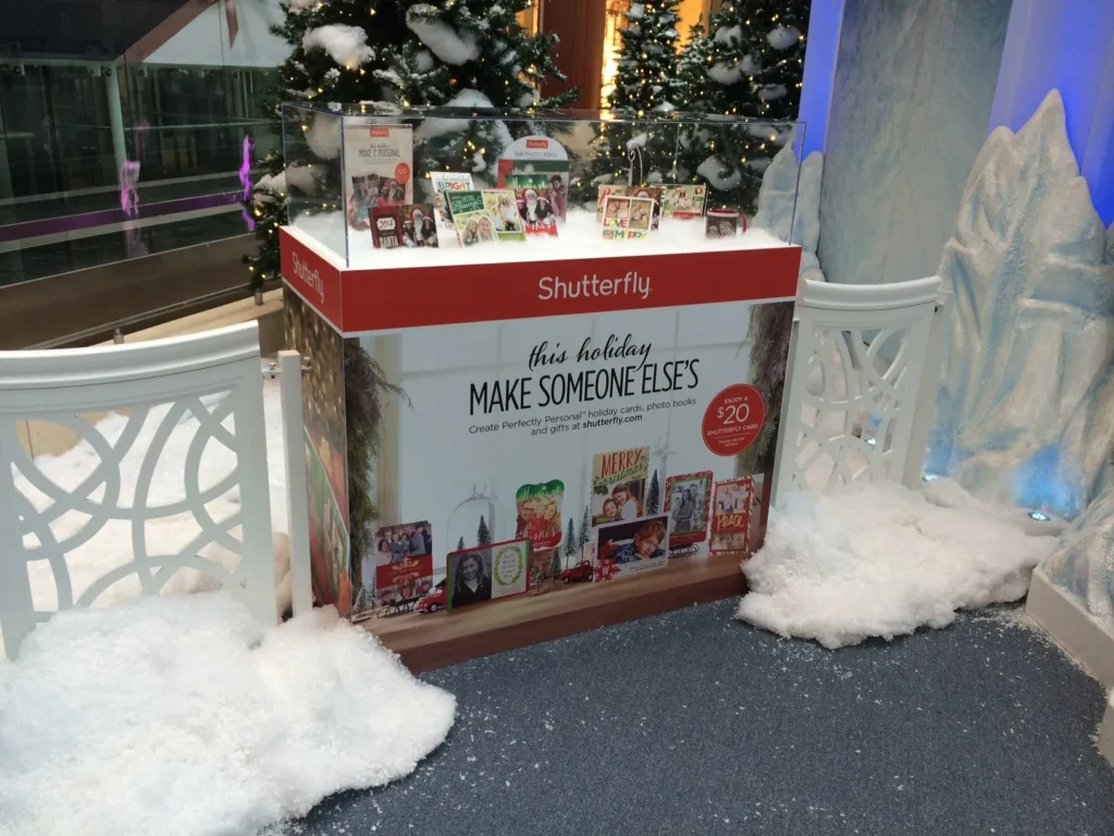 Shutterfly holiday display with photo gifts and promotional sign, surrounded by artificial snow, white chairs, and decorated trees.