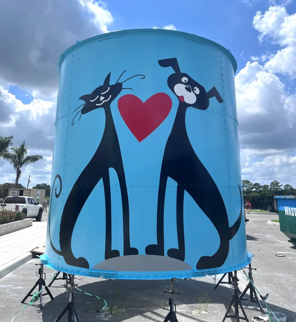Large blue tank features a cartoon painting of a black cat and black dog with a red heart between them, set outdoors under a partly cloudy sky.