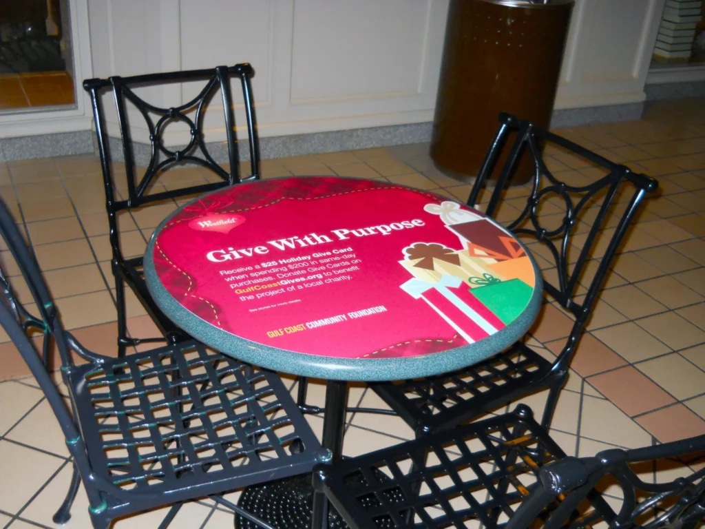 A round table with a “Give With Purpose” holiday-themed sign, surrounded by three black metal chairs, is set on a tiled floor indoors.