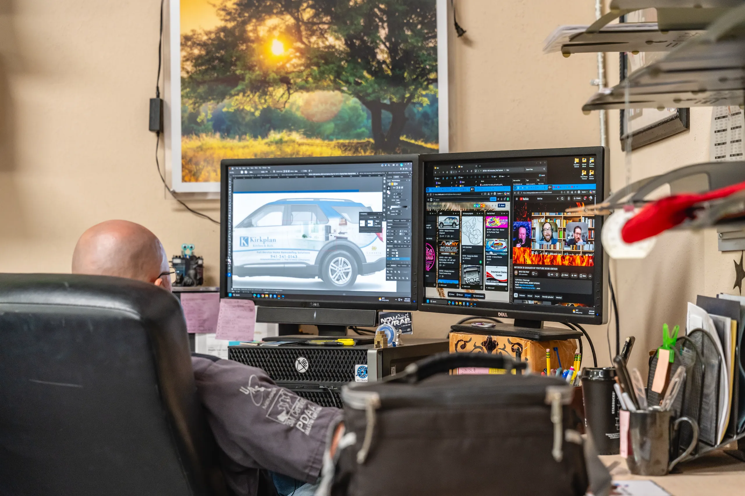 Person at desk working on two monitors; one shows a vehicle design, the other displays web pages; office setting with photo of a tree on the wall.