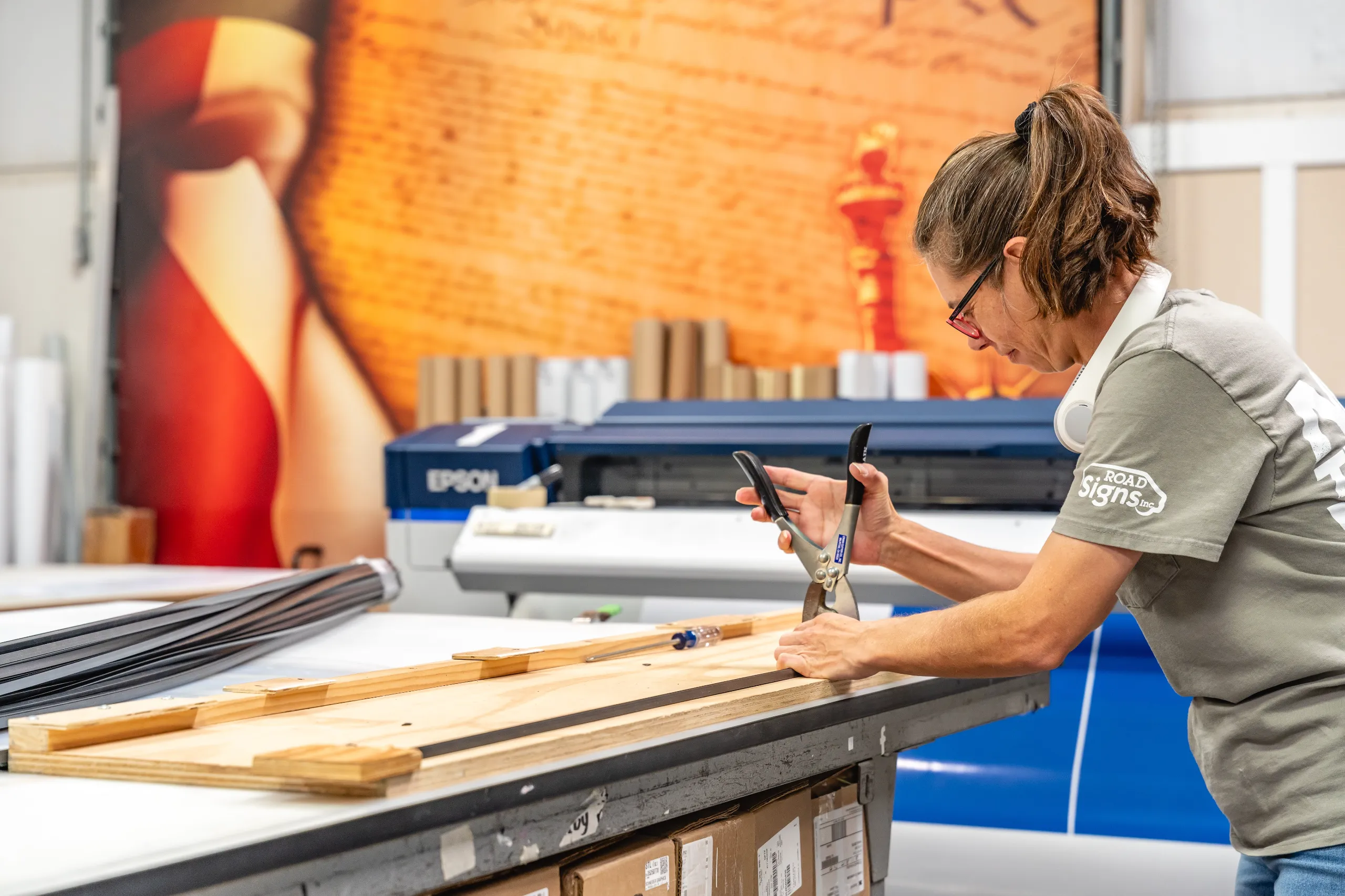 A woman uses a tool to cut or shape material on a work table in a print shop, with large equipment and a printer in the background.