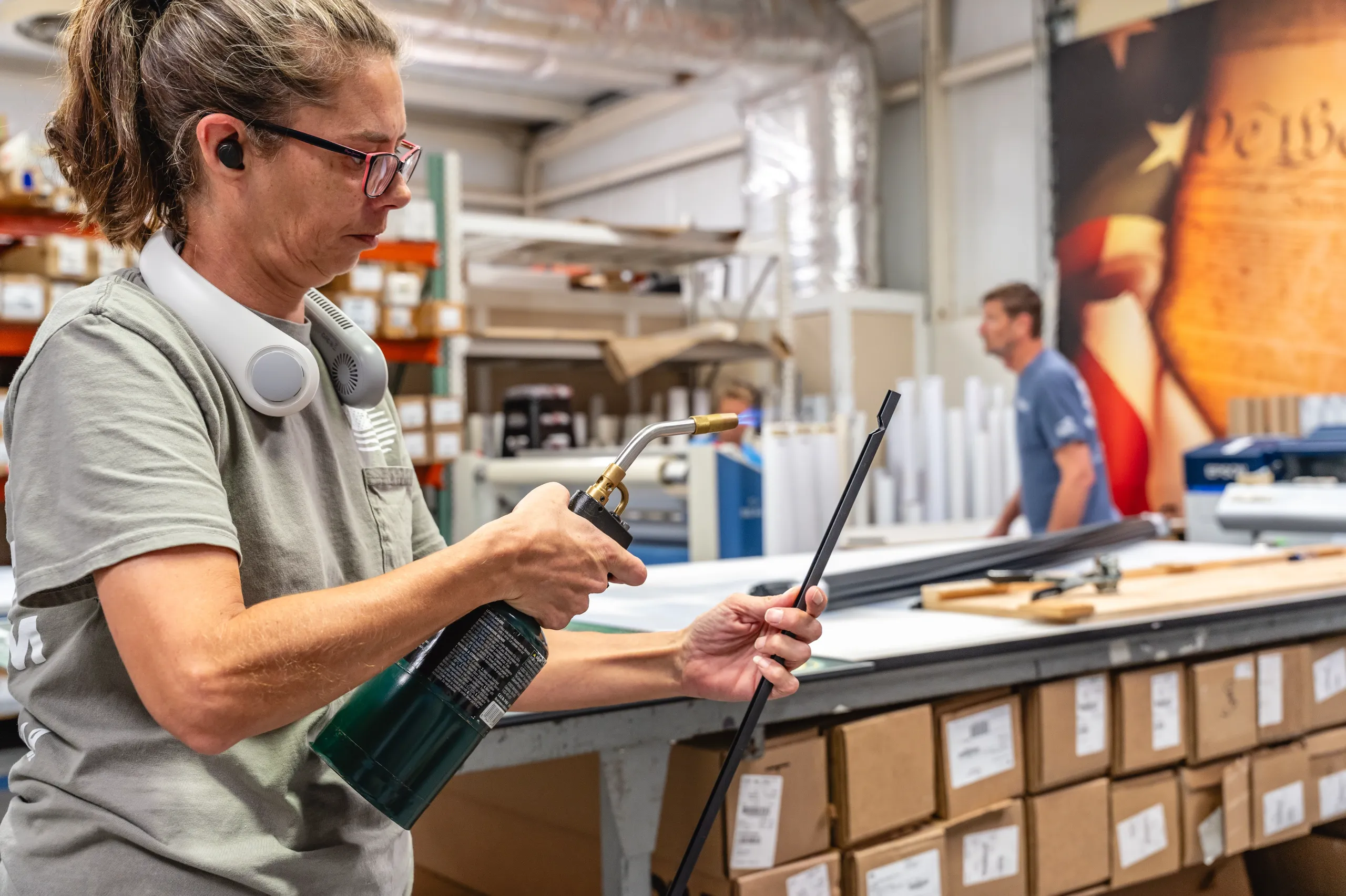 A woman wearing safety glasses uses a blowtorch on a metal rod in a workshop with boxes and two workers visible.