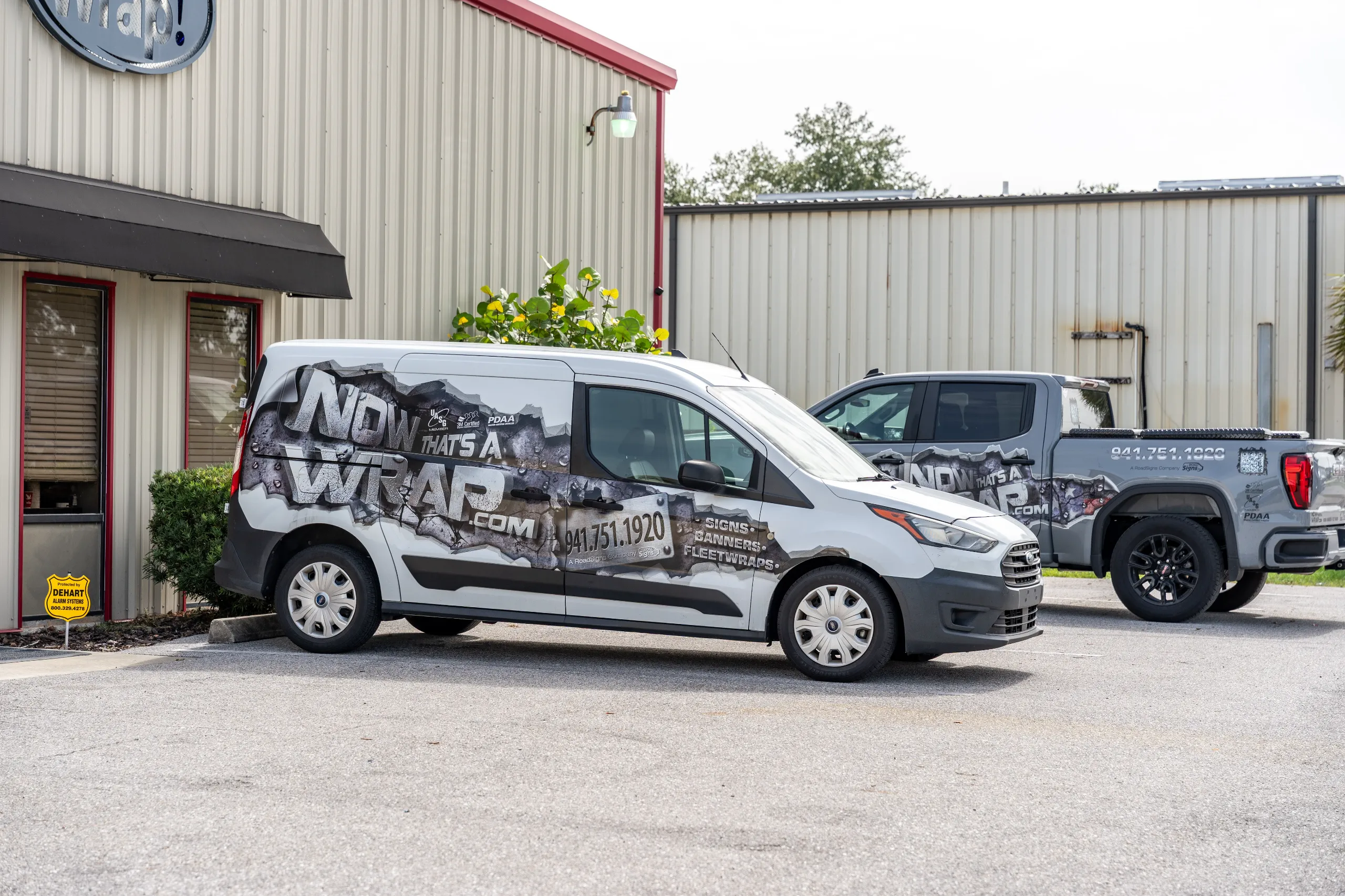 Two grey Now That’s A Wrap company vehicles with branded graphics are parked outside a building with metal siding and a red awning.