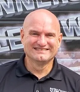 A smiling bald man in a black collared shirt stands in front of a wall with partially visible text in the background.