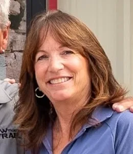 Woman with long brown hair and a blue shirt smiling at the camera, standing next to another person whose face is partially out of frame.