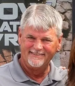 Smiling older man with gray hair and a goatee, wearing a gray collared shirt, standing in front of a textured wall with partially visible text.