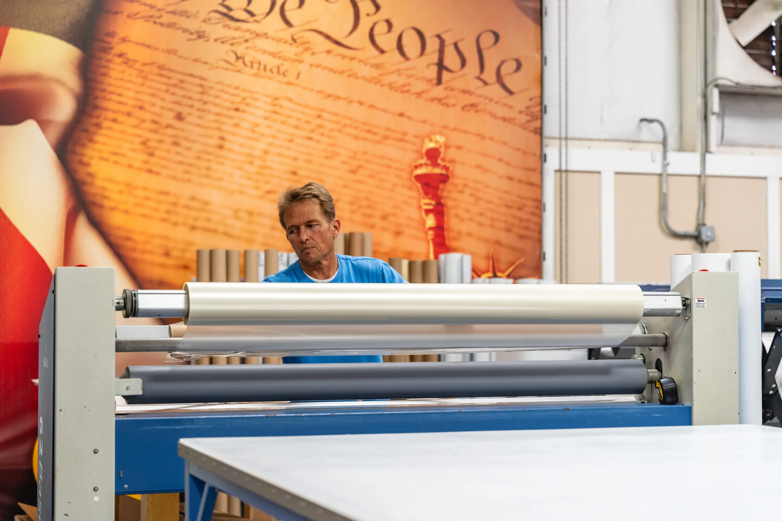 A man operates a large printing or laminating machine in front of a mural featuring the U.S. Constitution and the Statue of Liberty.