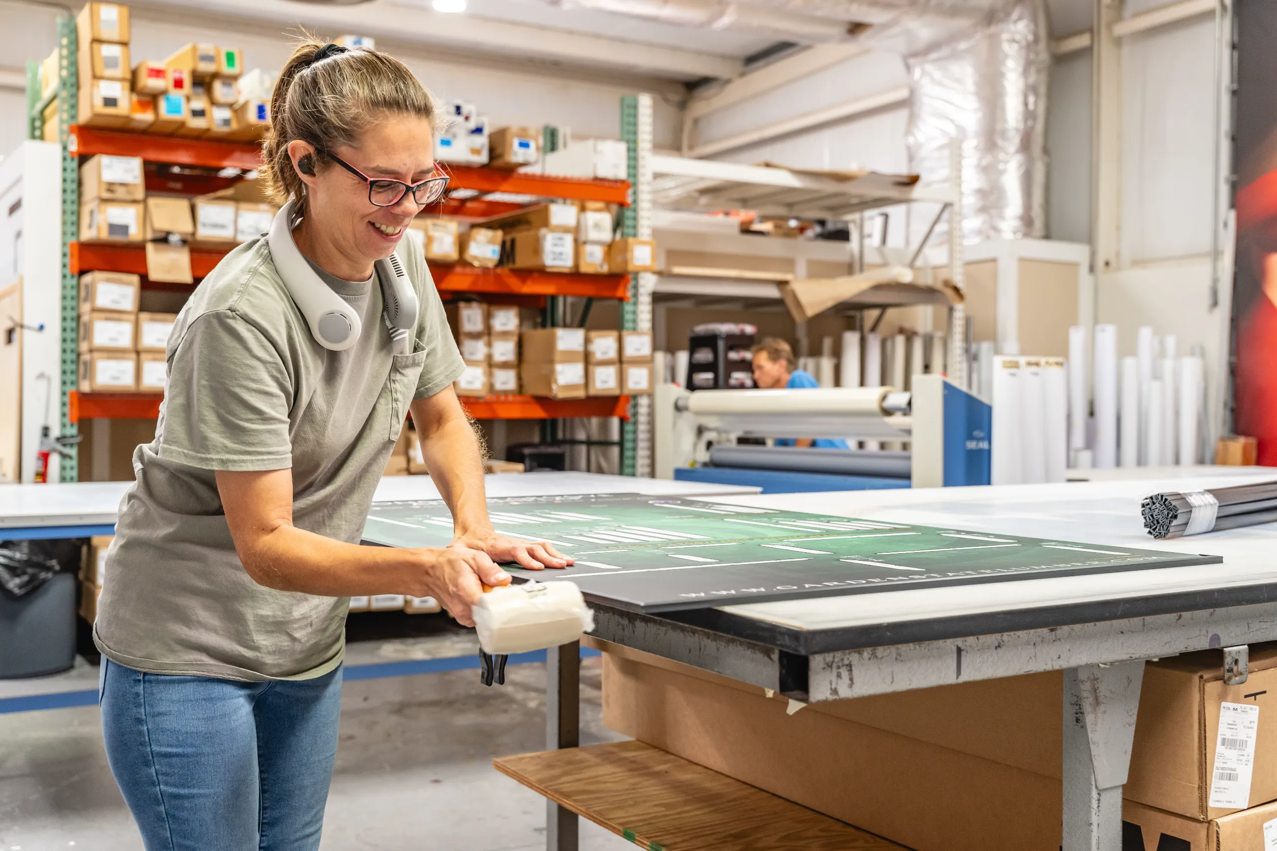 A woman in a workshop smooths a green printed sheet on a worktable, with shelves of boxes and industrial equipment in the background.